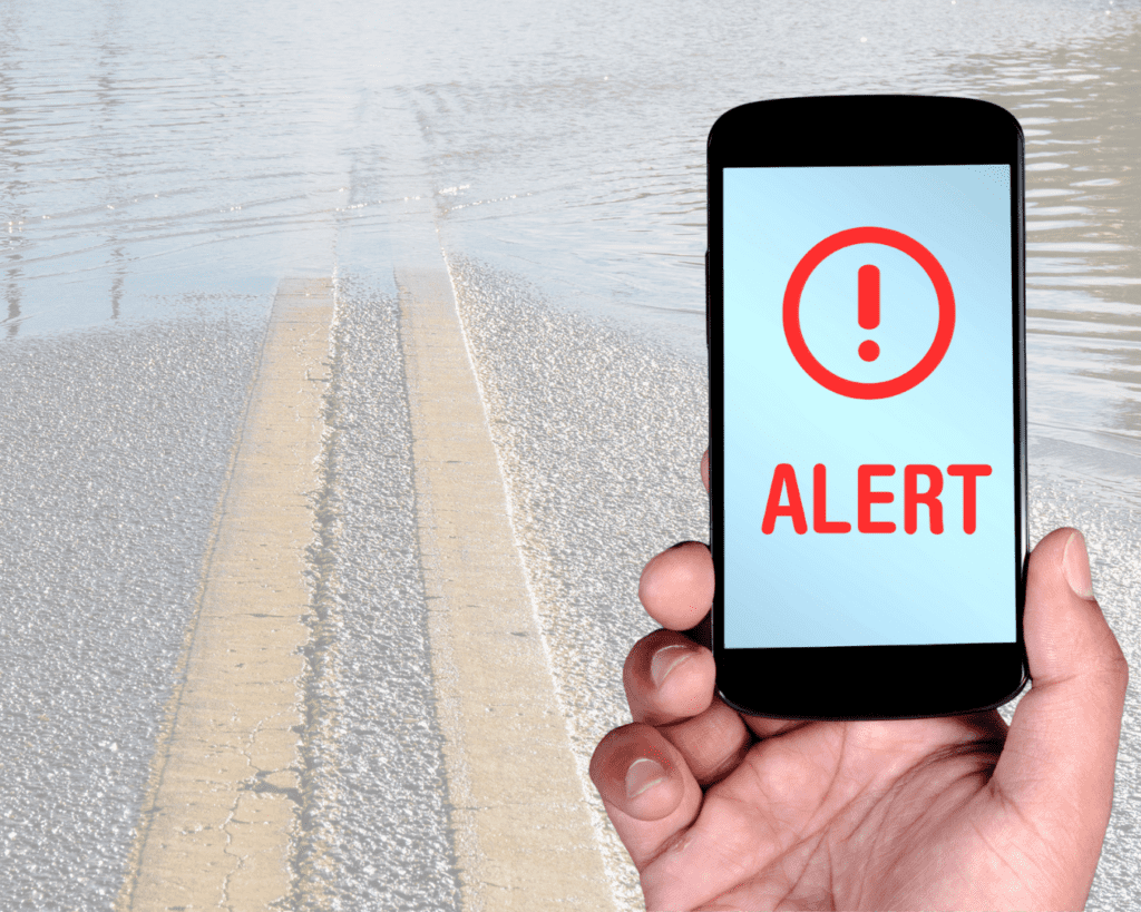 Flooded road with a hand holding a smartphone with the text Alert on it