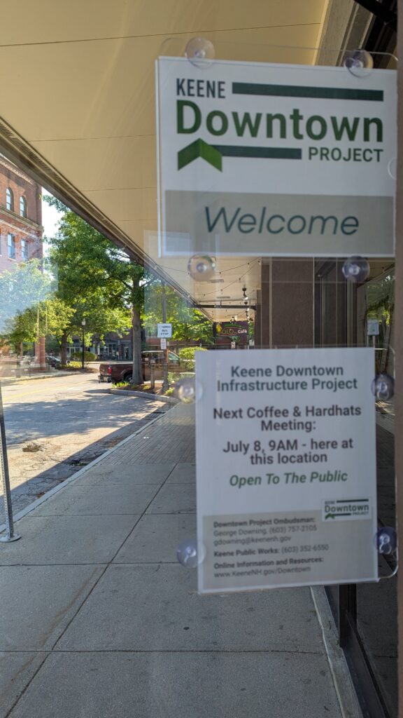 A small sign for the Keene Downtown Project  on an open door with a Keene street in the background